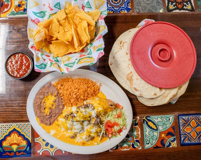 Cheesy beef entree with rice, refried beans, tortilla chips, salsa, and warm tortillas on a vibrant tiled table.