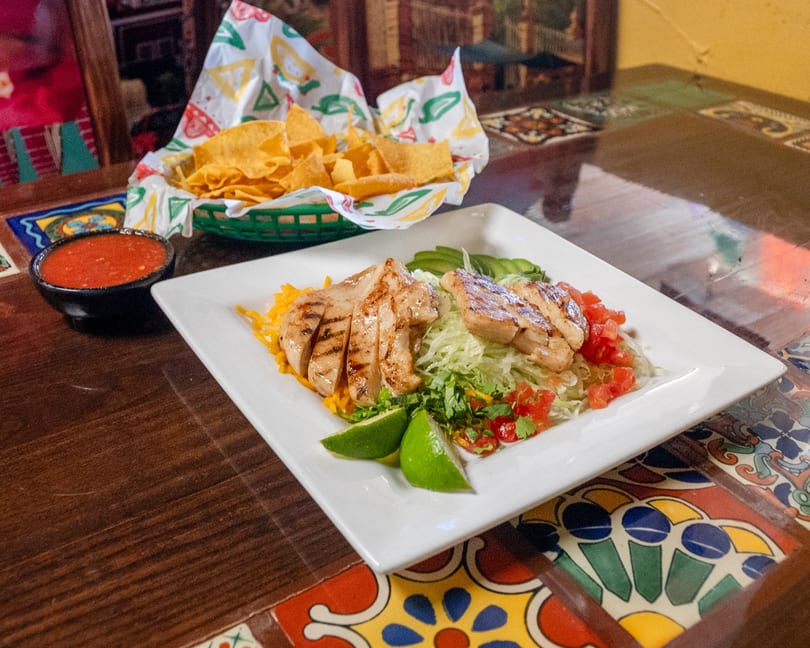 Grilled chicken salad with avocado, lime, and salsa, served with tortilla chips on a tiled table.