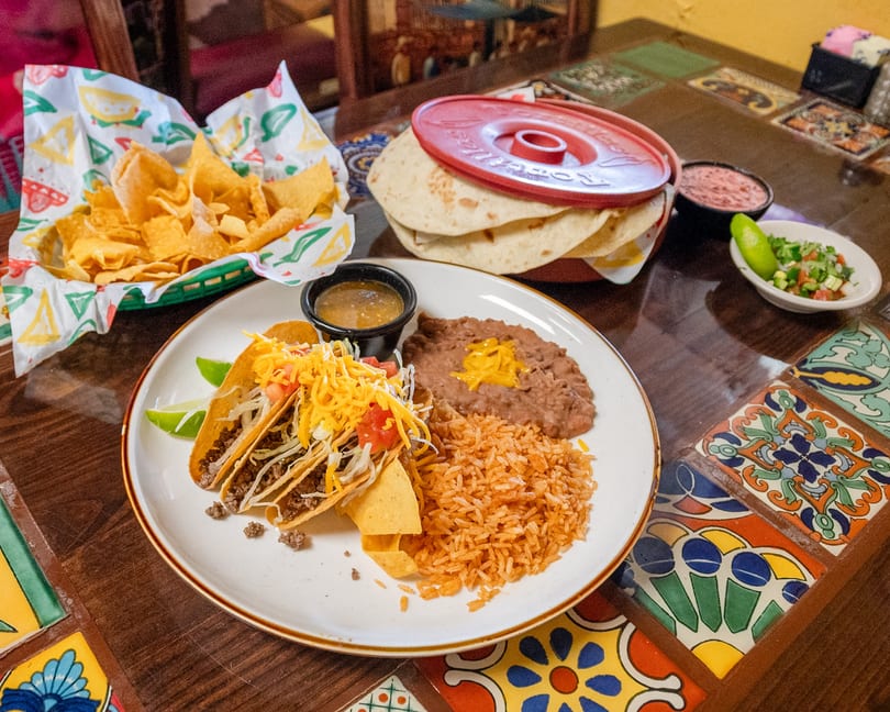 Mexican meal with three tacos, rice, refried beans, chips, and tortillas on a vibrant tiled table.