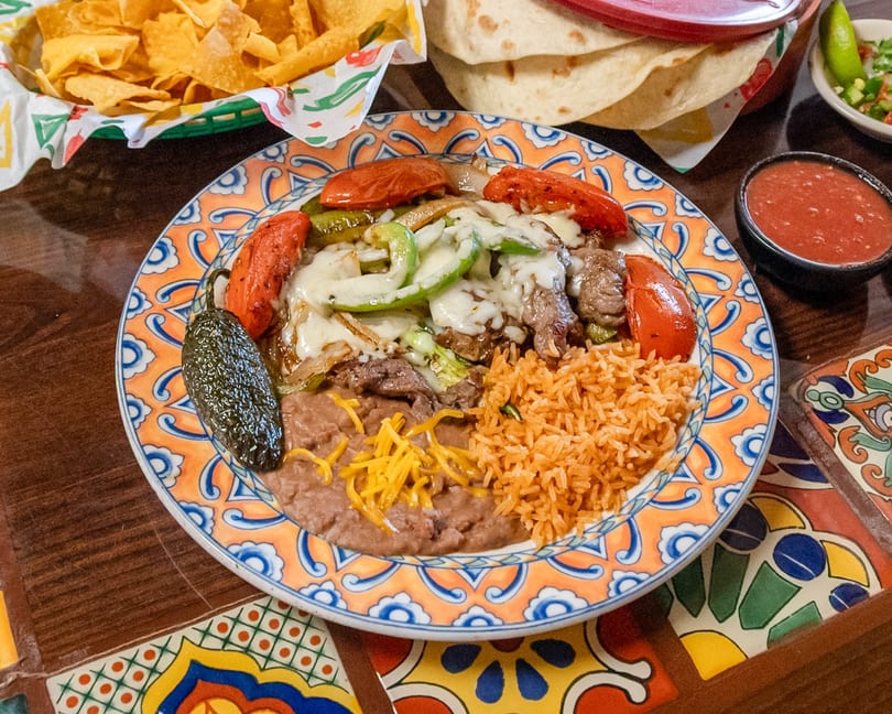 A vibrant Mexican fajita plate featuring steak, melted cheese, bell peppers, rice, beans, and a jalapeño.