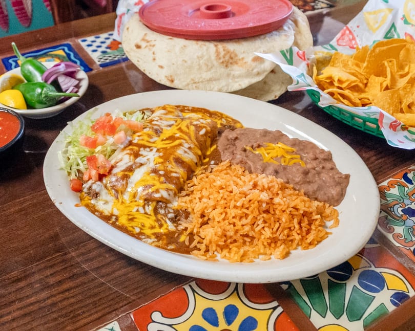 Mexican dinner plate with cheesy enchiladas, rice, beans, tortillas, chips, and salsa on a vibrant tiled table.