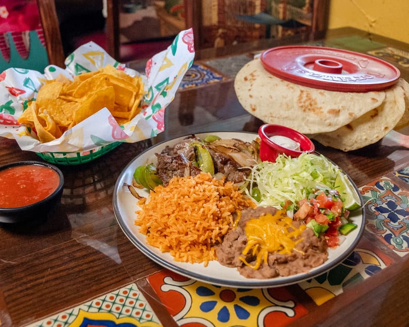 Fajita plate featuring steak, peppers, onions, rice, refried beans, lettuce, pico de gallo, chips, salsa, and tortillas.