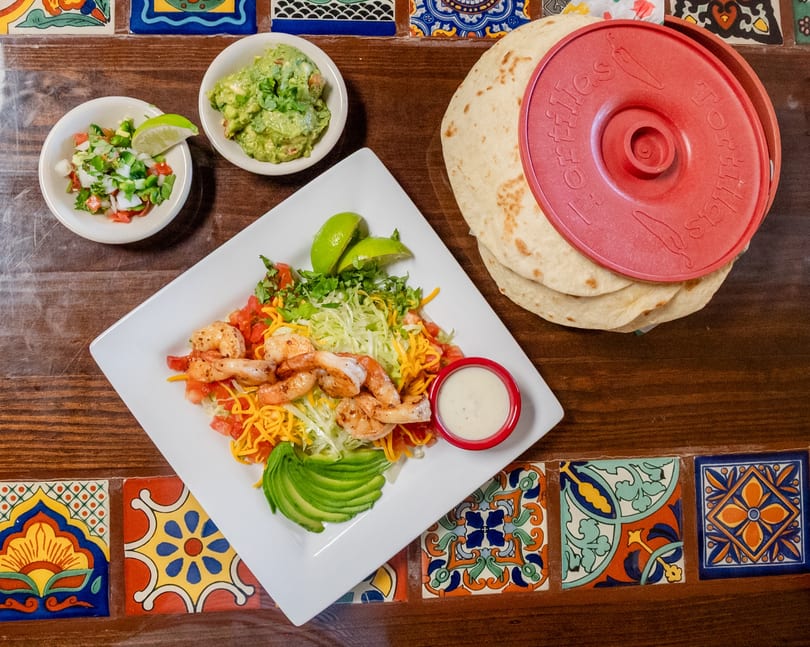 Grilled shrimp fajita plate with guacamole, pico de gallo, and tortillas on a tiled wooden table.