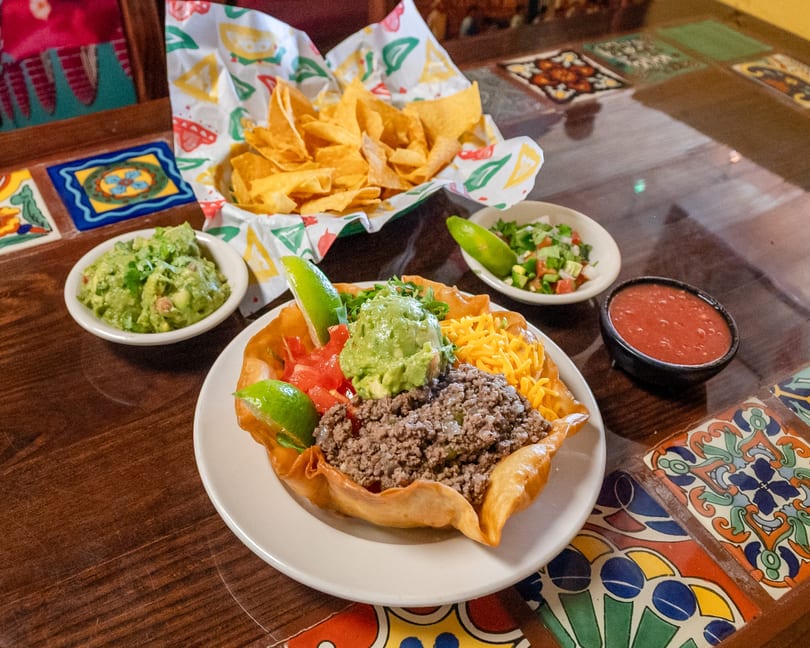 Colorful taco salad with ground beef, guacamole, and cheese, alongside chips, salsa, and pico de gallo on a tiled table.