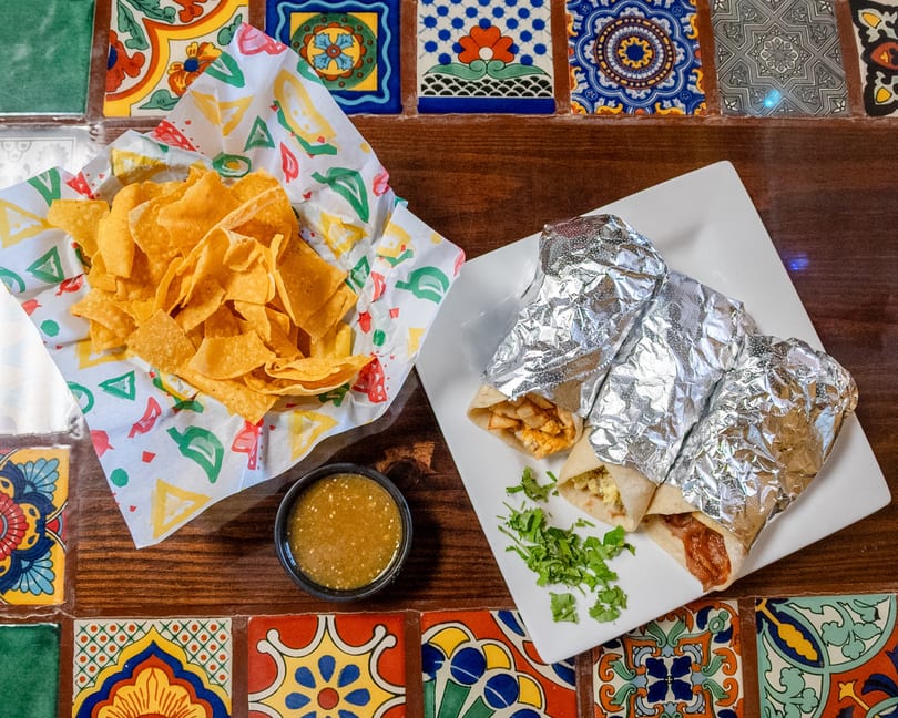 Three burritos, a basket of tortilla chips, and green salsa on a colorful tiled table.