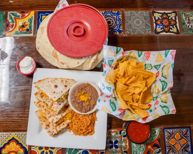 Quesadillas with rice, beans, chips, salsa, sour cream, and tortillas on a colorful tiled table.